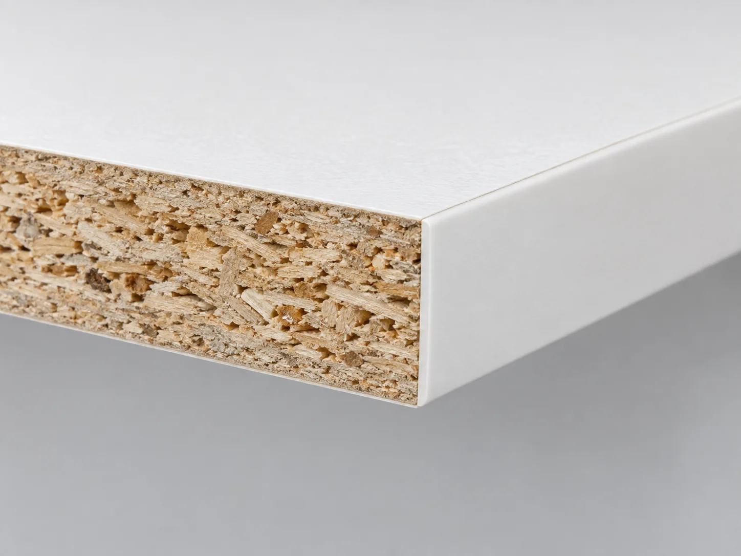 Cut corner of a white melamine panel showing the thin fused decorative surface over a particleboard core, with white edge banding on the finished edge